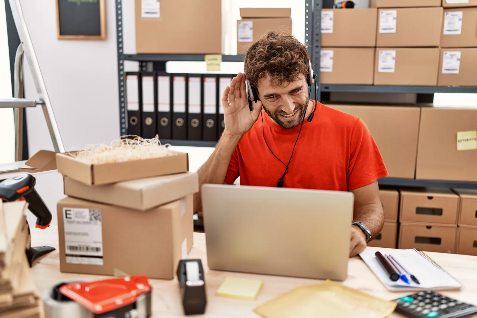 Young hispanic call center agent man working warehouse smiling with hand ear listening hearing rumor gossip deafness concept